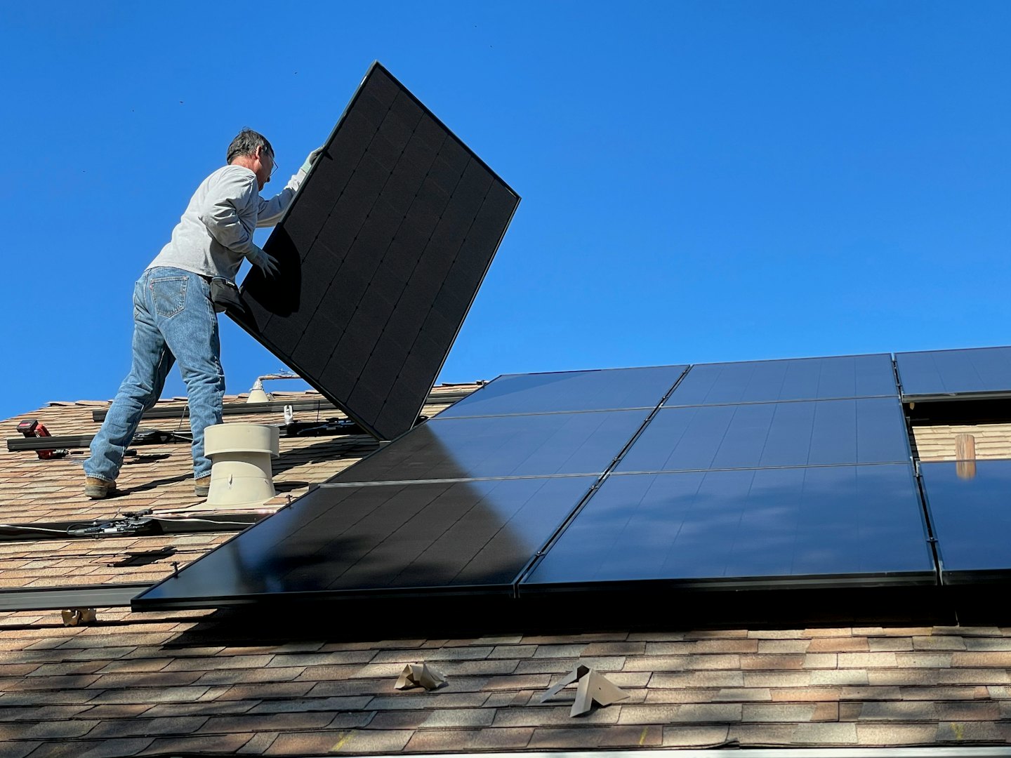 Technician installs solar panel on shingled roof under blue sky.
